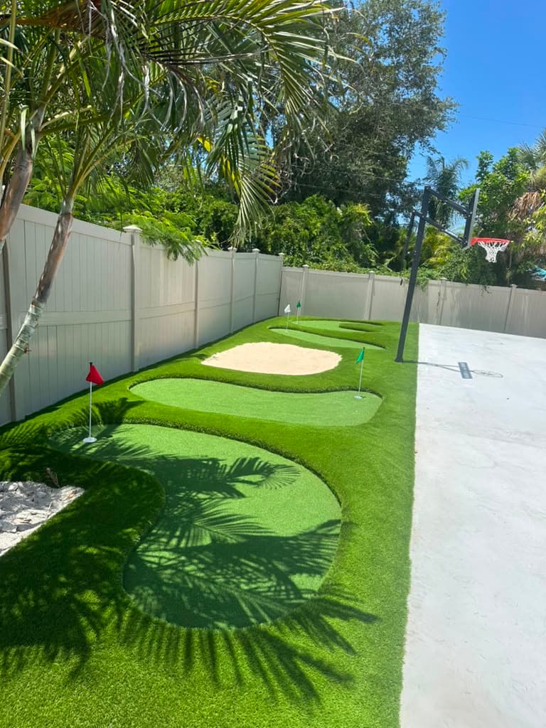 Backyard putting green built from artificial turf with sand bunker and flags, framed by palm trees